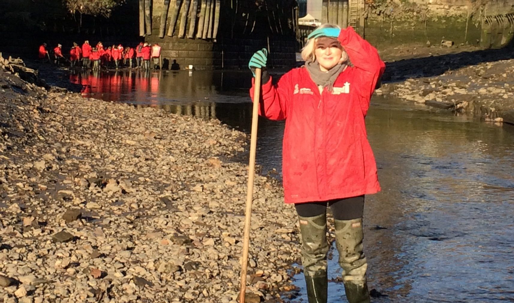 a photo of ella frears, a woman in a bright read coat with short very blonde hair, standing in a creek leaning on a large supporting stick