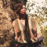 a person with long dark hair wearing a muted stripy top leans against a vast, old tree trunk