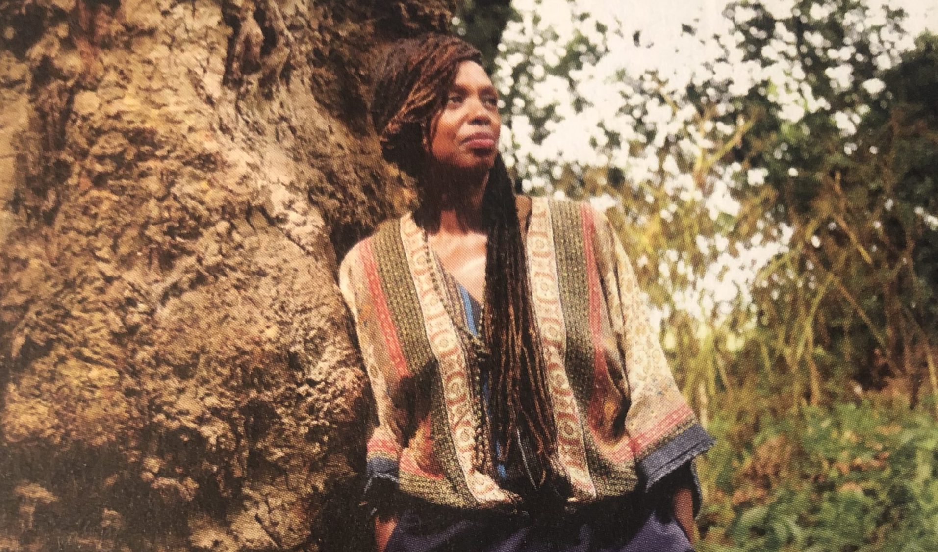 a person with long dark hair wearing a muted stripy top leans against a vast, old tree trunk