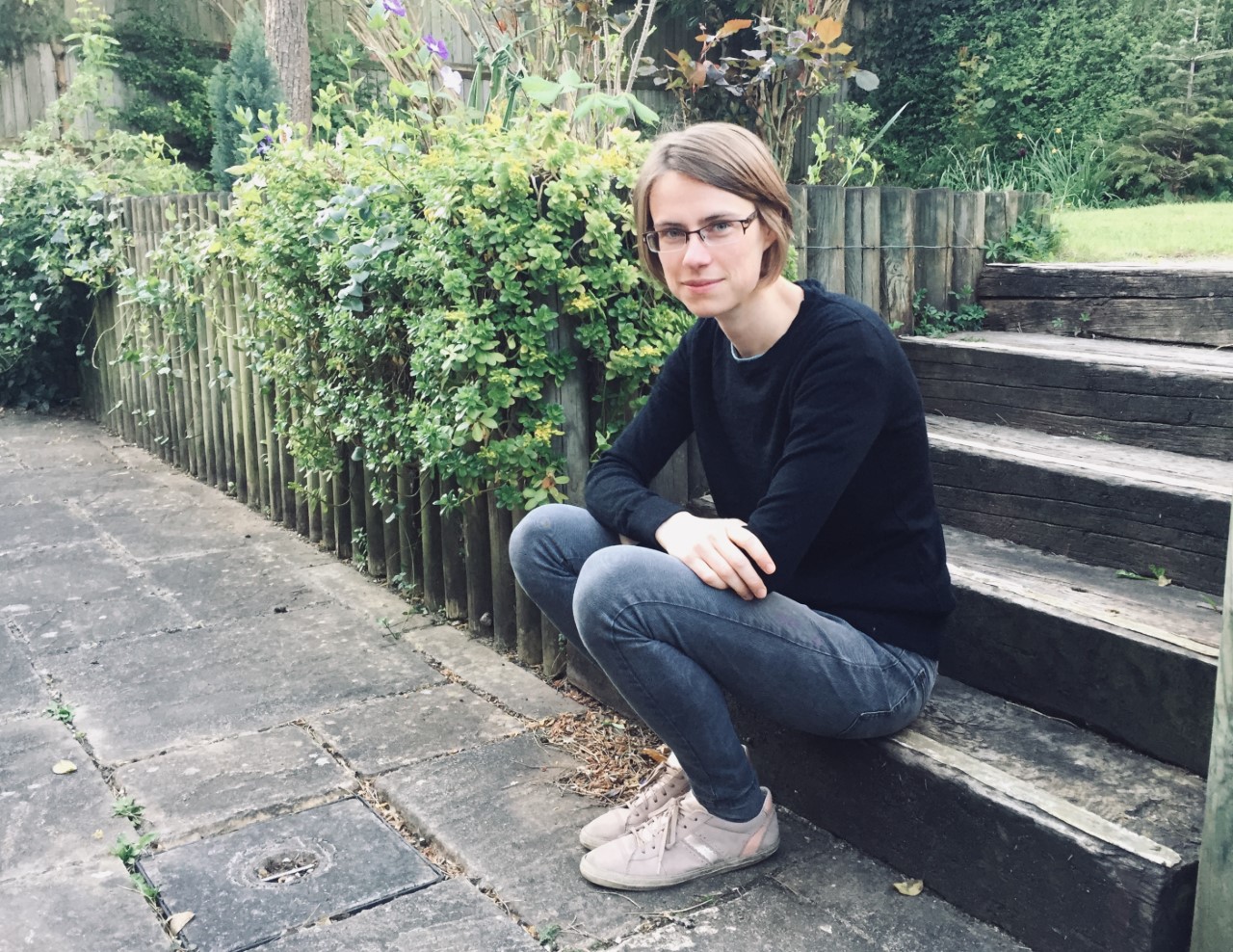 A woman with short dark blonde hair wearing glasses, a blue jumper and blue jeans sitting on some steps looking at the camera.