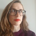 a woman with long brunette hair looking up to the right. She's wearing red lipstick and big black glasses. 