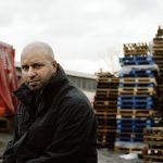 A picture of Amer Anwar, wearing a black jacket and looking into the camera. In the background are part of a lorry, high stacks of empty wooden pallets and a grey sky.