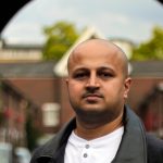 a picture of a man standing inbetween a row of buildings looking thoughtfully at the camera