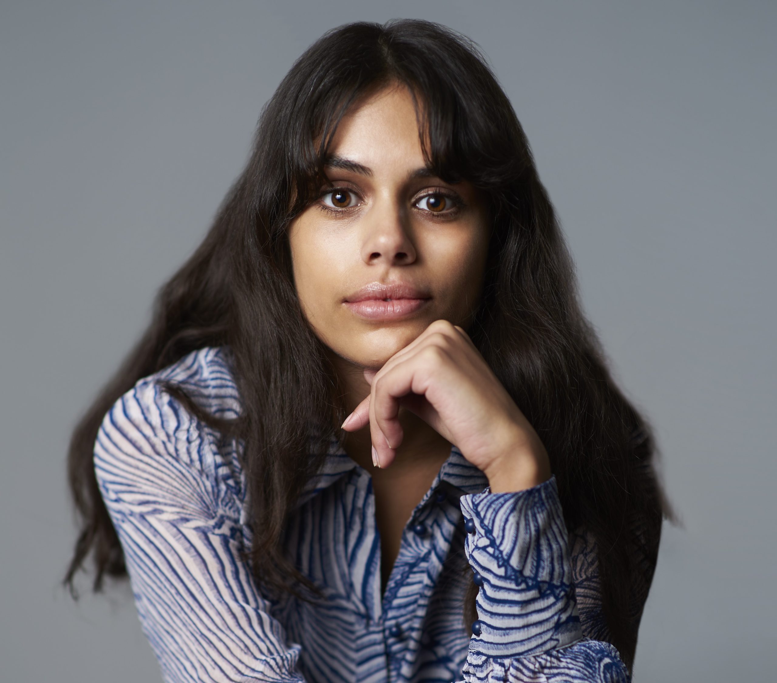 Headshot of a woman looking directly at the camera with her chin resting on her hand. She is shown against a pale grey background. She has long wavy dark brown hair falling over her shoulders and is wearing a long-sleeved blue and white stripe-patterened shirt.