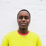 a black woman with cropped hair wearing a bright yellow jumper standing against a white wall