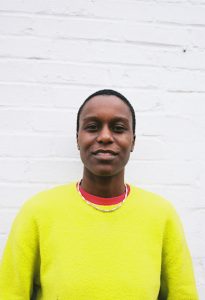 a black woman with cropped hair wearing a bright yellow jumper standing against a white wall