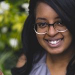 The image is a close up of a Black woman with shoulder length black hair wearing brown and gold glasses. She is wearing a sleeveless grey top, looking directly at the camera and smiling. Behind her is a blurred image of flowers and foliage.