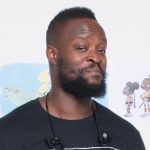 a black man in a Basquiat TShirt smiles at the camera in front of a white wall with illustrations of characters