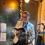 Writer Chris Simpson reading at a live event - he is standing in front of a microphone stand and reading from a book