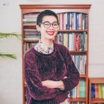 Helen Bowell, a Chinese and British young woman, wearing a purple jumper smiles in front of a bookcase.