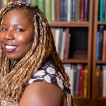 Maame Blue smiling and looking over her left shoulder with a book case behind her