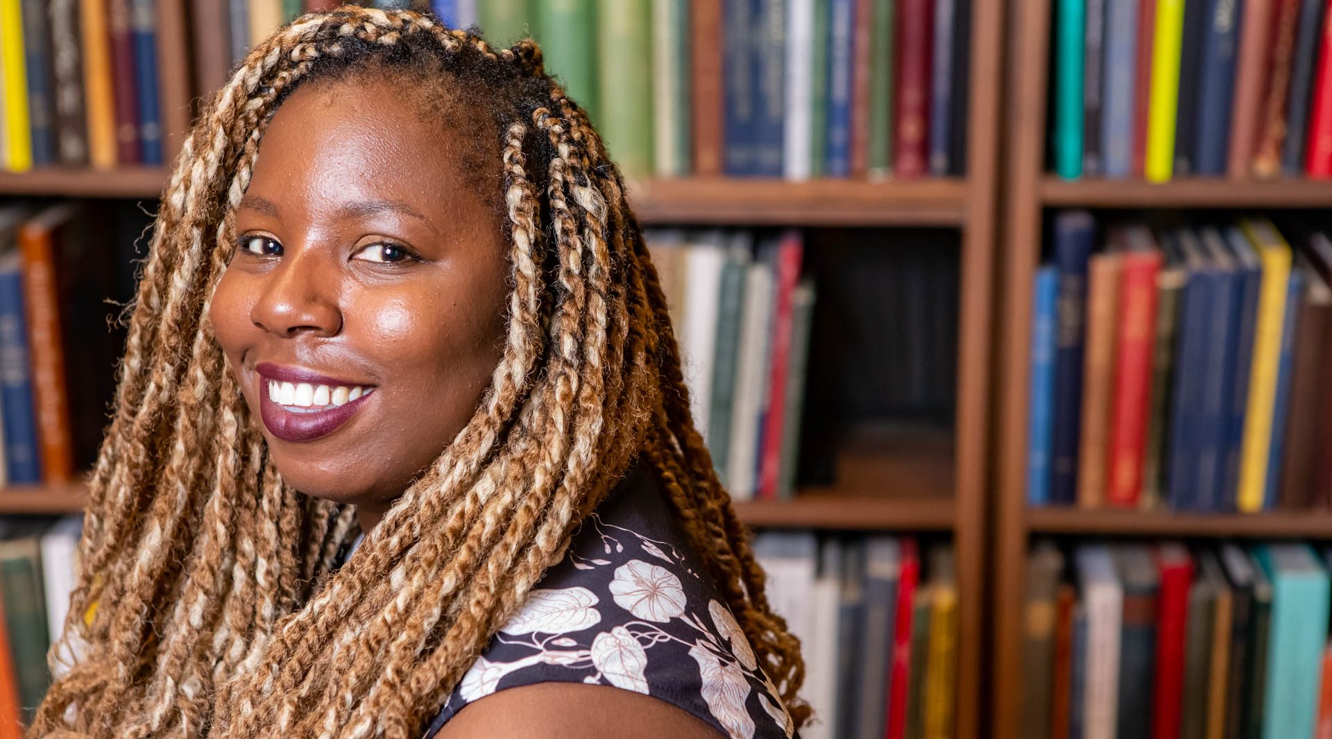 Maame Blue smiling and looking over her left shoulder with a book case behind her