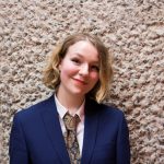 The image shows a white person, Annie Hayter, smiling. They have bobbed blondish hair, black winged eyeliner and red lipstick. They are wearing a navy-blue suit jacket with a white shirt and paisley patterned tie, and are standing up against a crunchy-looking cement wall, face on to the camera.