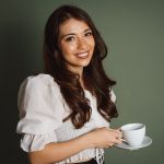 a photo of a woman wearing a frilly white shirt, holding a teacup and saucer. She has long wavy brunette hair. 