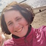 a woman with short dark bobbed hair smiling at the camera. She is on a beach, which you can see in the background.