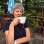 a woman with short curly grey hair sat at a table drinking from a white mug