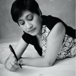 Black and white photo of an Indonesian woman with short hair, earrings, and a patterned dress, lying down on her front, pen in hand, ready to write. Picture credit: Derrick Kakembo.
