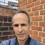 a photo of a man with short cropped hair standing in front of a brick wall