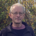 a man standing in front of some trees. He wears glasses and is wearing a dark blue shirt. 