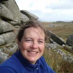 a woman sitting in front of some rocks. She is smiling widely and is wearing a blue jumper.