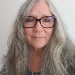 a woman with long grey hair and black glasses. She looks thoughtfully at the camera. 