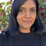 a woman wearing a black jumper in front of a raspberry bush. She has shoulder length black hair and is looking thoughtful.