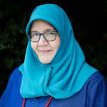 a woman wearing a bright blue hijab. She is wearing glasses and looks thoughtfully at the camera.