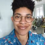 a photo of a woman with short dark hair and big glasses. She is smiling widely and wearing a blue patterned shirt.