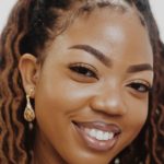 a closeup of a woman smiling widely. She is wearing dangly gold earrings and her hair is in twists and is partially tied up. 