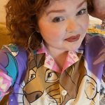 a person with shoulder length brown/ read curly hair wearing a colourful shirt with a tiger on it