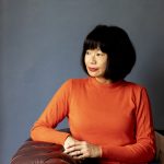 a woman with a short sleek dark blob wearing an orange jumper. She is sat at a table and looks over to her side