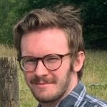 a photo of a man wearing glasses looking at the camera. He is in the countryside.