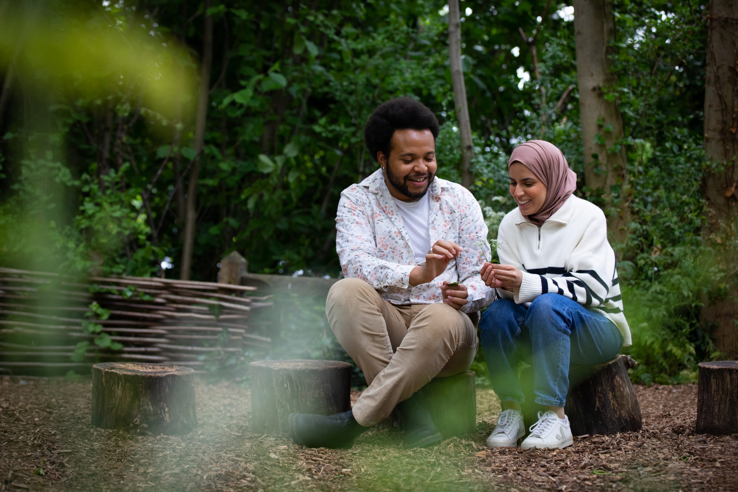 an image of two people sitting on a bench in a wooded area looking at small plants.