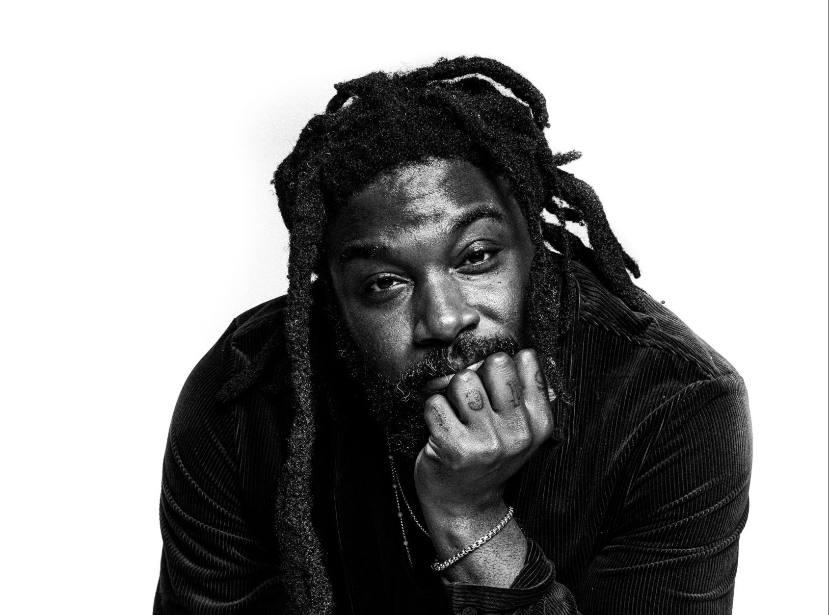 a photo of a Black man with dreadlocks sitting on a chair, with his head leaning on his hand in a thoughtful pose