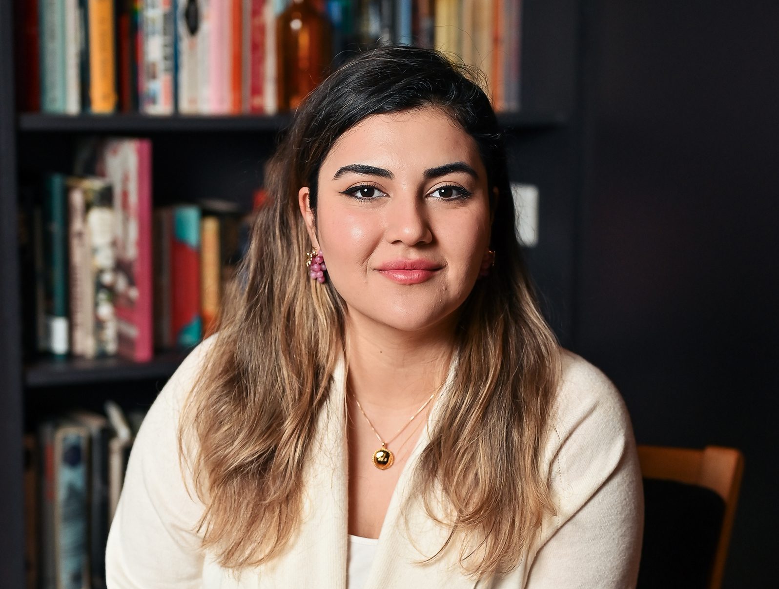 a person with long hair, dark brown at the roots and lighter brown at the ends, sits on a chair in front of a tall bookcase. They are dressed smartly and smiling.