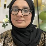 Sahera has light brown skin, a round face, round black glasses, black headscarf, and a black dress with a gold pattern. She is smiling slightly. The background is blurred green, gold and white decorations.