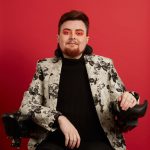 Jamie Hale, a white person with red hair and beard looks directly towards the camera with a confident smile. They sit in their electric wheelchair in front of vivid red/orange photographers banner, wearing black jeans, a ribbed black turtleneck and a silver floral blazer. A shock of bright pink and orange eyeshadow brings out their green eyes.