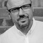 The photograph shows a middle-aged white man wearing a white shirt. He has glasses, is bald and has a short greying beard. (The image is by Naomi Woddis).