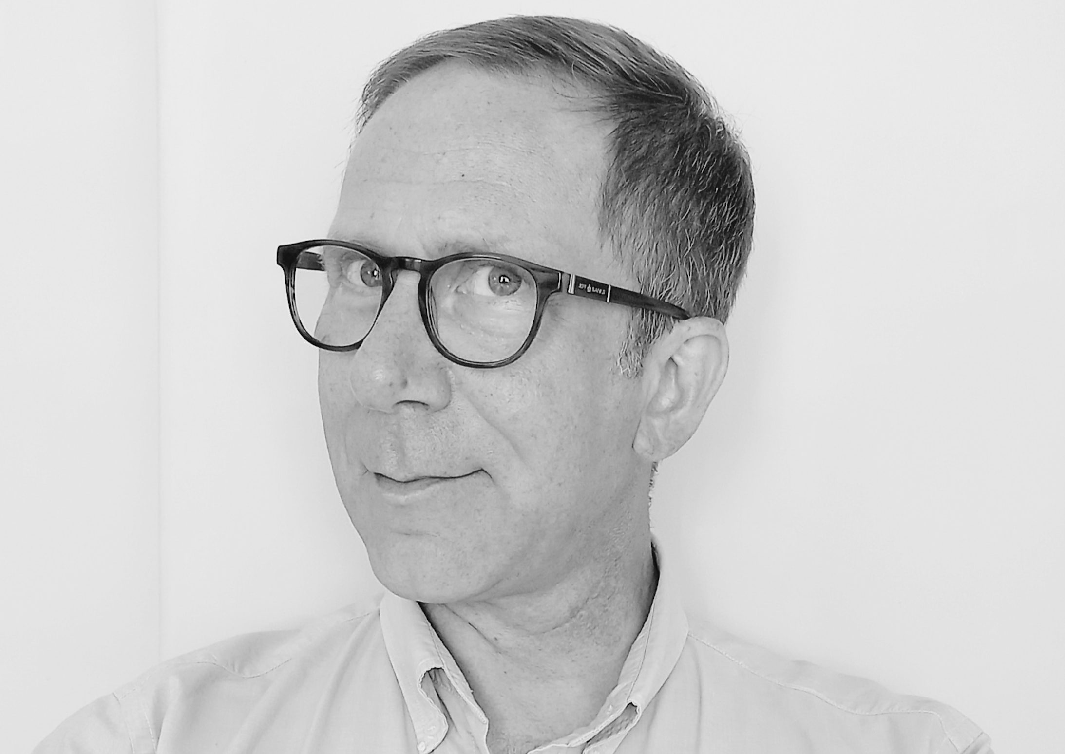 Black and white image of man with fair hair wearing glasses and a white shirt turning his head to the side and smiling.