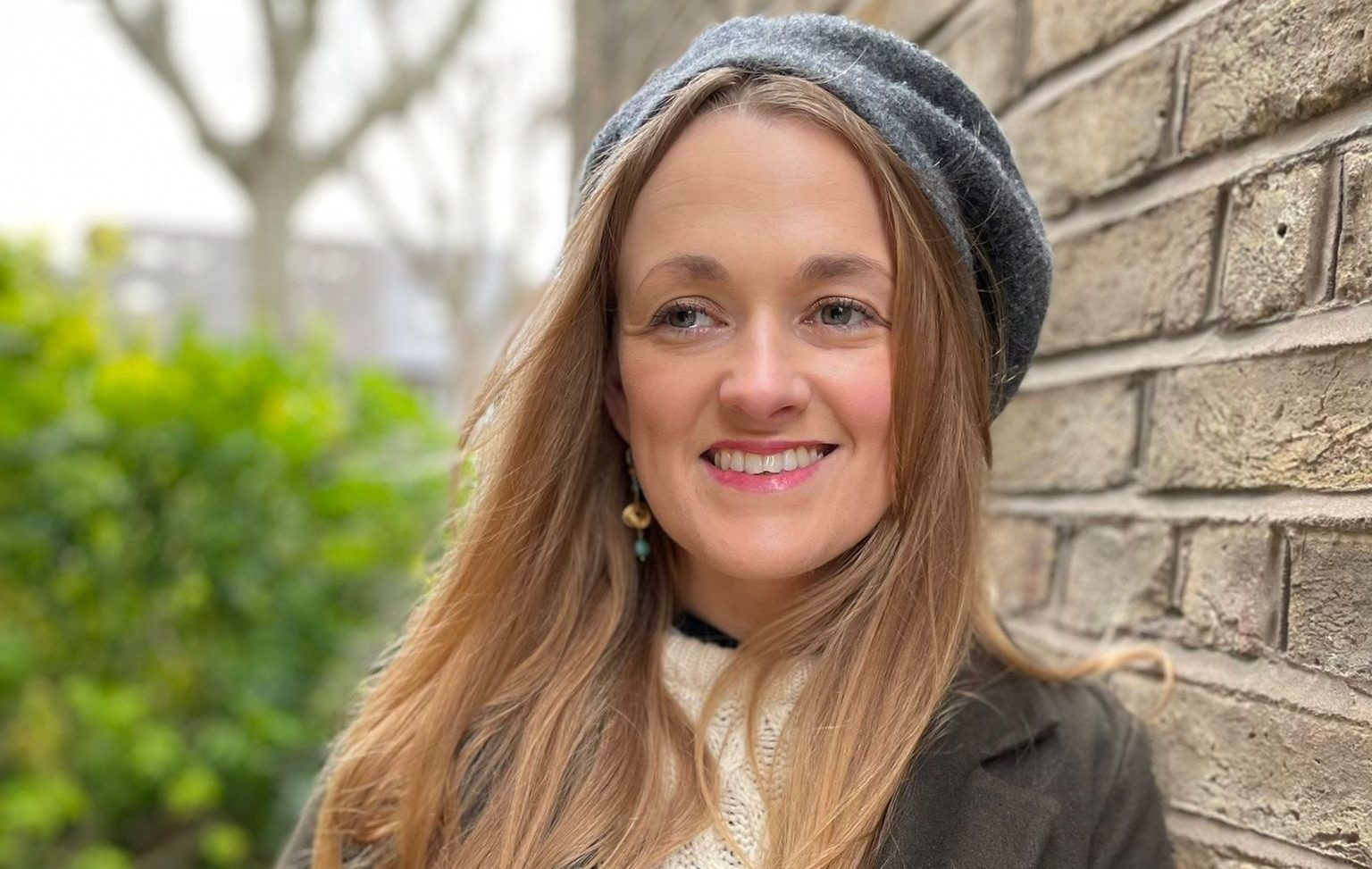 A woman with long light brown hair smiles wearing a grey hat, a white long sleeved top and dark grey blazer