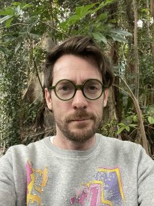 Adam Zmith is a man with glasses and a beard taking a selfie in a forest 