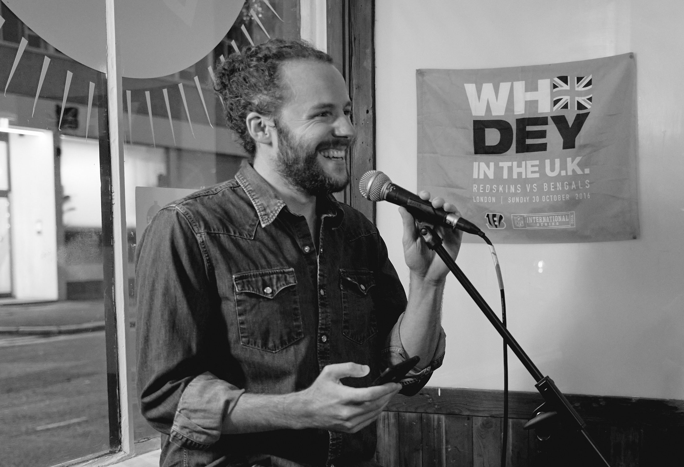 A man with dark, curly hair and a beard smiles widely looking to the side with one hand poised on a microphone.