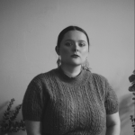 A black and white photo of a person sat in a room with a blank wall behind them. 