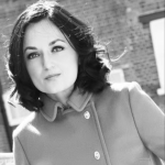 Black and white photo of a person smiling in the street, wearing a buttoned up coat.