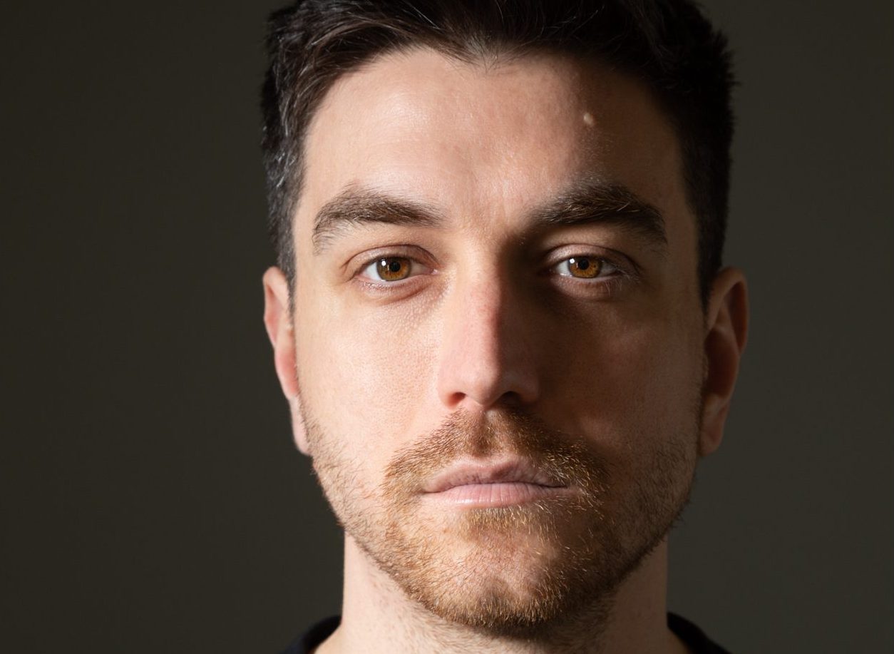 A man with dark hair wearing a black jumper stares forward. The background behind him is dark grey.