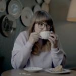 A photo of Laurie in a lilac sweatshirt drinking tea from a white/floral cup & saucer