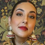 A woman wearing large earrings with a colourful patterned shirt in front of colourful wallpaper