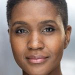 a black woman with short cropped hair wearing a grey / blue jumper smiles at the camera