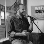 A bearded, smiling white man in his mid-thirties wearing a denim shirt and jeans. He sits in front of a microphone and holds a mobile phone.