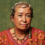 Pascale is wearing an embroidered orange top and an orange and green necklace, she has silver braided hair and is looking straight at the camera.